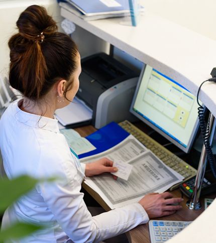 A Medical Admin Assistant with brown hair in a bun sits at a desk, working on a computer. She holds papers, including a medical form, and is surrounded by office supplies, folders, and a printer.