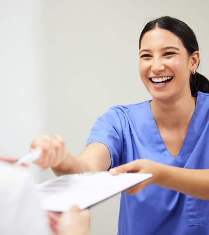 A smiling healthcare worker in blue scrubs, possibly an LPN, hands a clipboard and pen to another person, suggesting a friendly interaction in a medical setting—a glimpse into the supportive environment of nursing jobs.