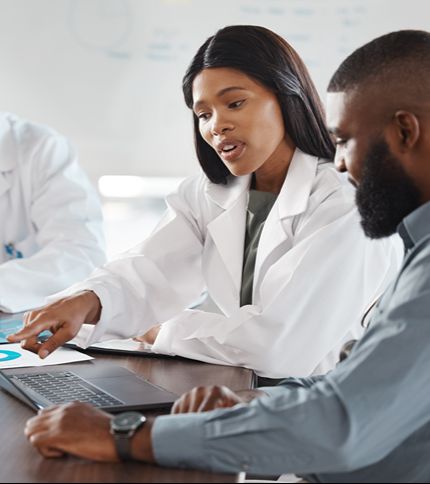 A Health Service Manager in a white lab coat points at a laptop screen while talking to a man beside her. Another medical professional is partially visible as they discuss something in a professional setting.
