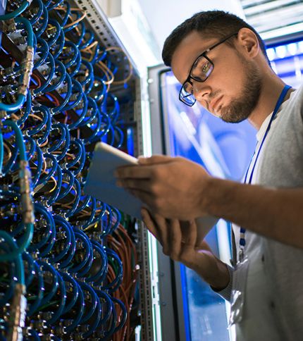 A Network Administrator wearing glasses uses a tablet while inspecting a server rack filled with cables and electronic equipment in a data center.