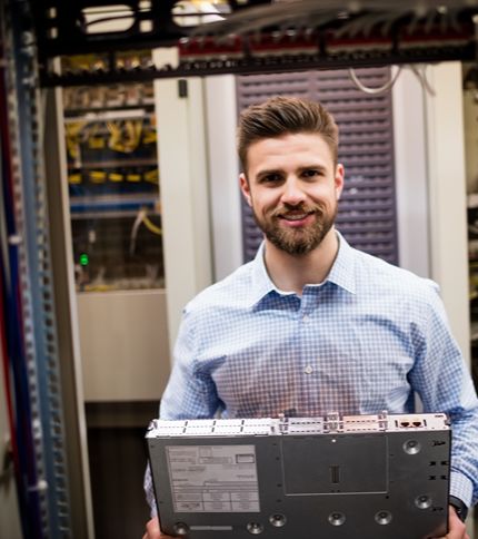 A bearded Network Technician in a checked shirt stands in a server room, holding server equipment and smiling at the camera.