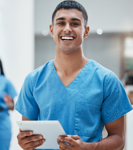 A smiling Nursing Assistant in blue scrubs holds a tablet in a brightly lit medical facility.