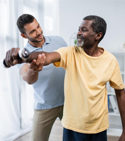 A Physical Therapist Assistant (PTA) assists an older man in a yellow shirt with an arm exercise, helping him lift a dumbbell in a bright, indoor setting. Both appear focused and engaged in the physical therapy activity.