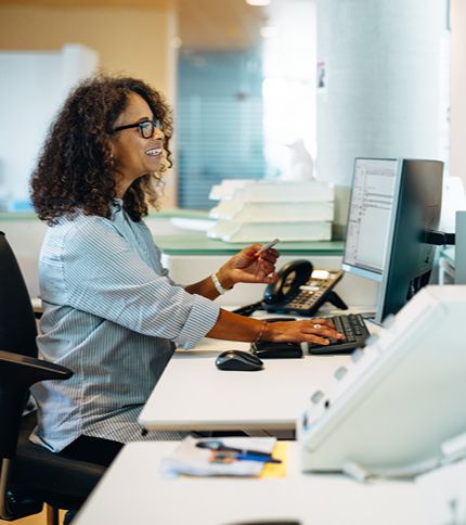 A smiling receptionist with curly hair and glasses sits at an office desk, working on a computer. She holds a pen in one hand, surrounded by office equipment and paperwork.