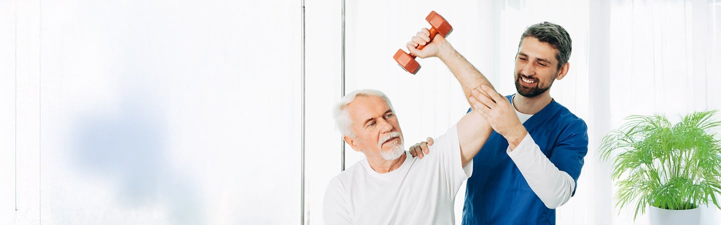 An Occupational Therapy Assistant helps an older man lift a small red dumbbell, guiding his arm during a rehabilitation exercise in a bright room with a plant in the background.