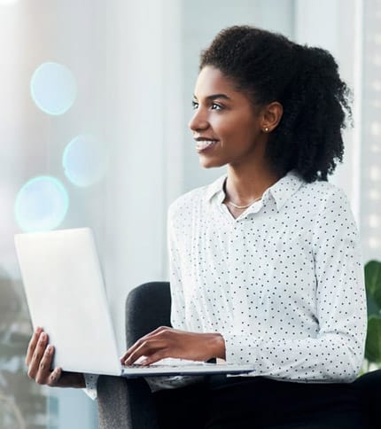 A woman with curly hair sits in an office chair smiling, holding a laptop on her lap, and looking to the side. She wears a white blouse with black polka dots. The background is softly blurred.