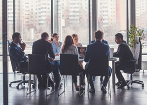 Eight people in business attire sit around a conference table in a modern office with large windows, having a meeting. City buildings are visible outside.