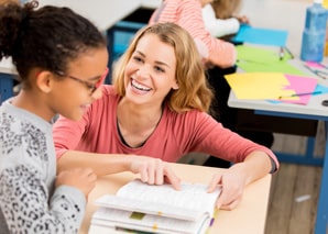 A smiling teacher sits at a desk, pointing to a book while talking with a young student who is wearing glasses. Another student is visible in the background, working with colorful papers.