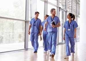 Four healthcare professionals in blue scrubs walk down a brightly lit hospital corridor, talking and holding clipboards and files. Large windows let in natural light.
