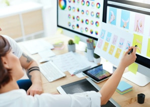 Two people working at a desk with computers, reviewing colorful graphic designs on the screen. One person points at the monitor with a stylus, surrounded by design tools and color palettes.