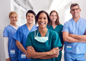 Five healthcare professionals in scrubs stand smiling in a hallway, with one woman in green scrubs and a surgical mask around her neck standing confidently at the front.