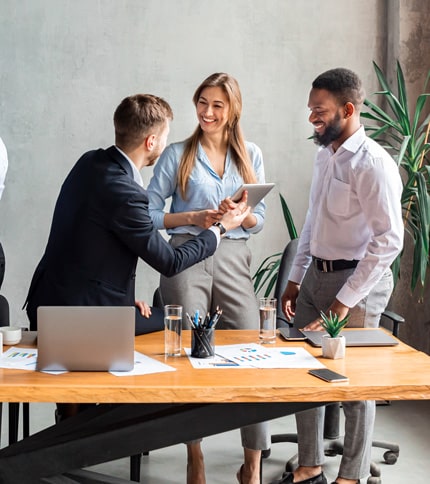 Three young professionals, two men and one woman, smile and interact around a modern office desk with laptops, papers, and plants, suggesting a collaborative and positive work environment.