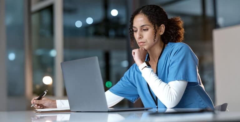 A healthcare worker in blue scrubs sits at a desk, looking at a laptop screen with a thoughtful expression while holding a pen, in a modern, well-lit office or hospital setting.