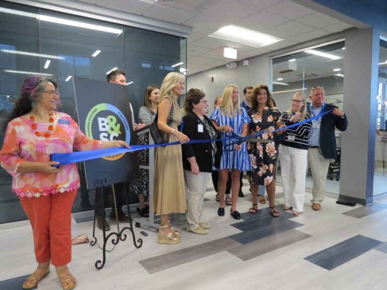 A group of people stand in a line indoors, smiling as they hold a large blue ribbon for a ribbon-cutting ceremony. The Bryant & Stratton College logo is displayed on an easel behind them at the Liverpool Campus Nursing Lab.