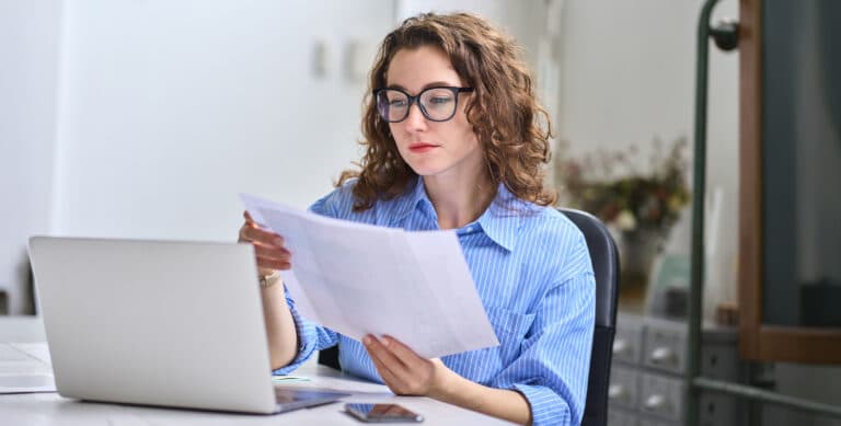 A woman with curly hair and glasses sits at a desk, reviewing medical billing papers in her hand while working on a laptop. A smartphone lies on the desk beside her in a bright, modern room.