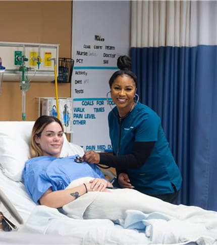 A nurse in blue scrubs smiles while using a stethoscope to check a patient in a simulated college experience, where students practice medical skills. The patient is covered with a blanket and looks relaxed, with medical equipment visible in the background.