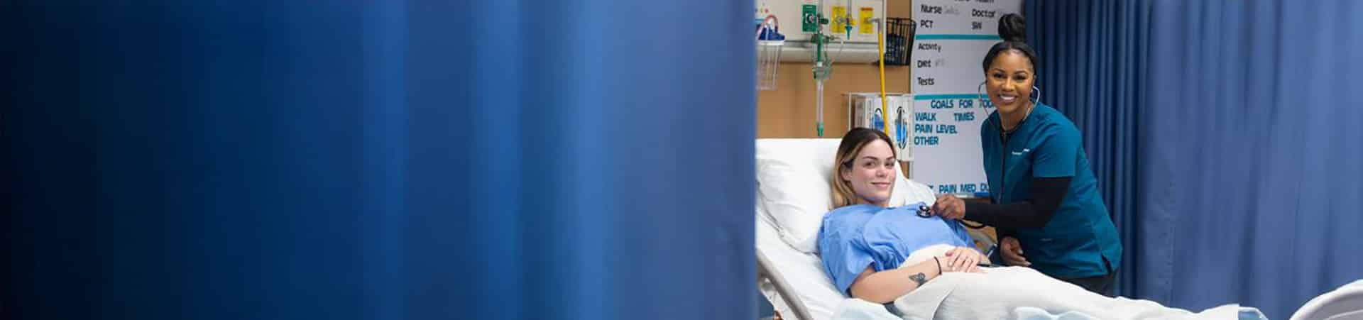 A nurse in blue scrubs smiles while checking a female patient's heartbeat with a stethoscope in a hospital bed, as part of a college simulation, separated by blue privacy curtains.