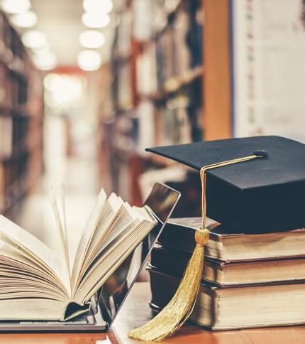 A graduation cap with a gold tassel rests on a stack of books beside an open book and a laptop on a library table, symbolizing the pursuit of knowledge and financial literacy, with bookshelves blurred in the background.