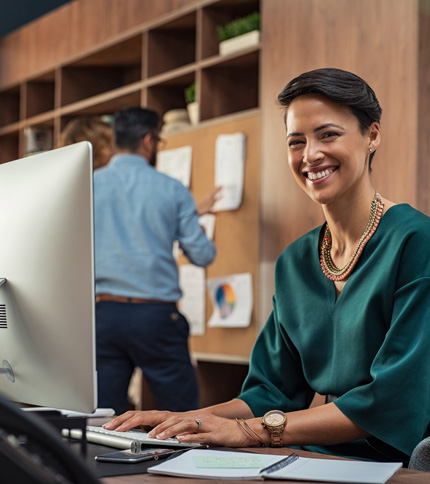 Smiling woman in a green blouse sits at her desk with a computer, while a man in the background pins papers to a corkboard—both engaged in tasks related to the Admissions Process in an office setting.