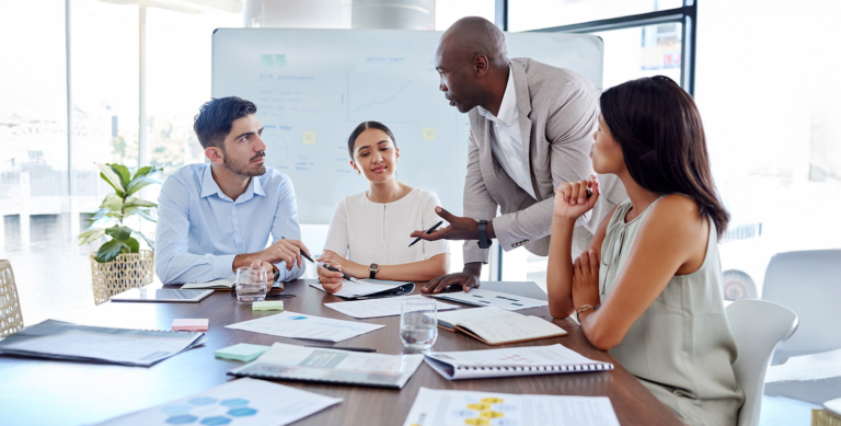 Four people in business attire sit around a table with papers and laptops, engaged in discussion. One person stands, gesturing while speaking, as the others listen attentively in a modern office setting.