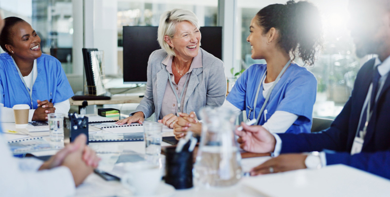 A group of healthcare professionals, including people in scrubs and business attire, sit around a conference table smiling and talking in a bright, modern office.