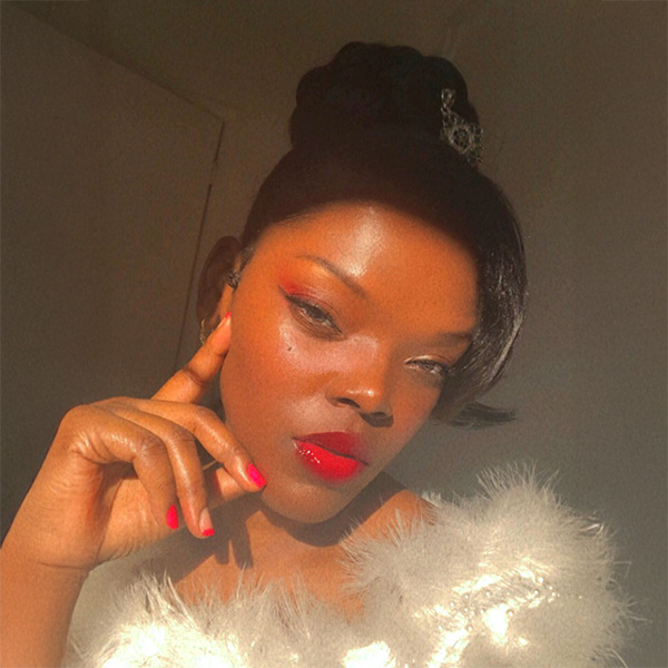 A woman with dark skin and red lipstick poses in soft sunlight, wearing a fluffy white garment. Her hair is styled up, and she touches her face gently, with red-painted nails and a calm, confident expression.