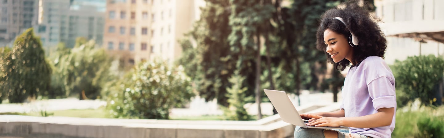 A young woman with curly hair, wearing headphones and a light purple shirt, sits outdoors on a ledge, smiling while using her laptop to explore Financial Aid Calculators. Trees and buildings are blurred in the background.