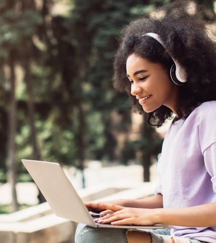 A young woman with curly hair, wearing headphones and a light purple shirt, sits outdoors smiling and typing on her laptop as she explores Financial Aid Calculators, with blurred trees in the background.