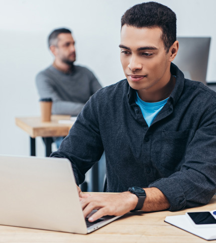 A young man wearing a dark shirt works on a laptop at a desk, possibly researching FAQs or terminology, while another person sits blurred in the background. A smartphone and coffee cup are on the desk.