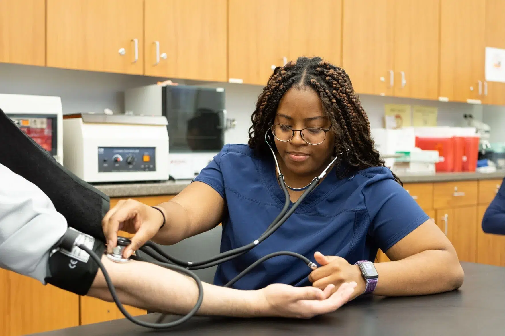 A healthcare professional in navy scrubs, measures a patient’s blood pressure with a stethoscope and cuff in a medical office with wooden cabinets in the background.