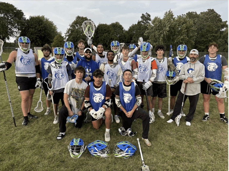 A group of Bryant & Stratton College men’s lacrosse players in blue and white uniforms pose together on a grassy field, most holding sticks and wearing helmets. Trees and a soccer goal are visible in the Wisconsin background.