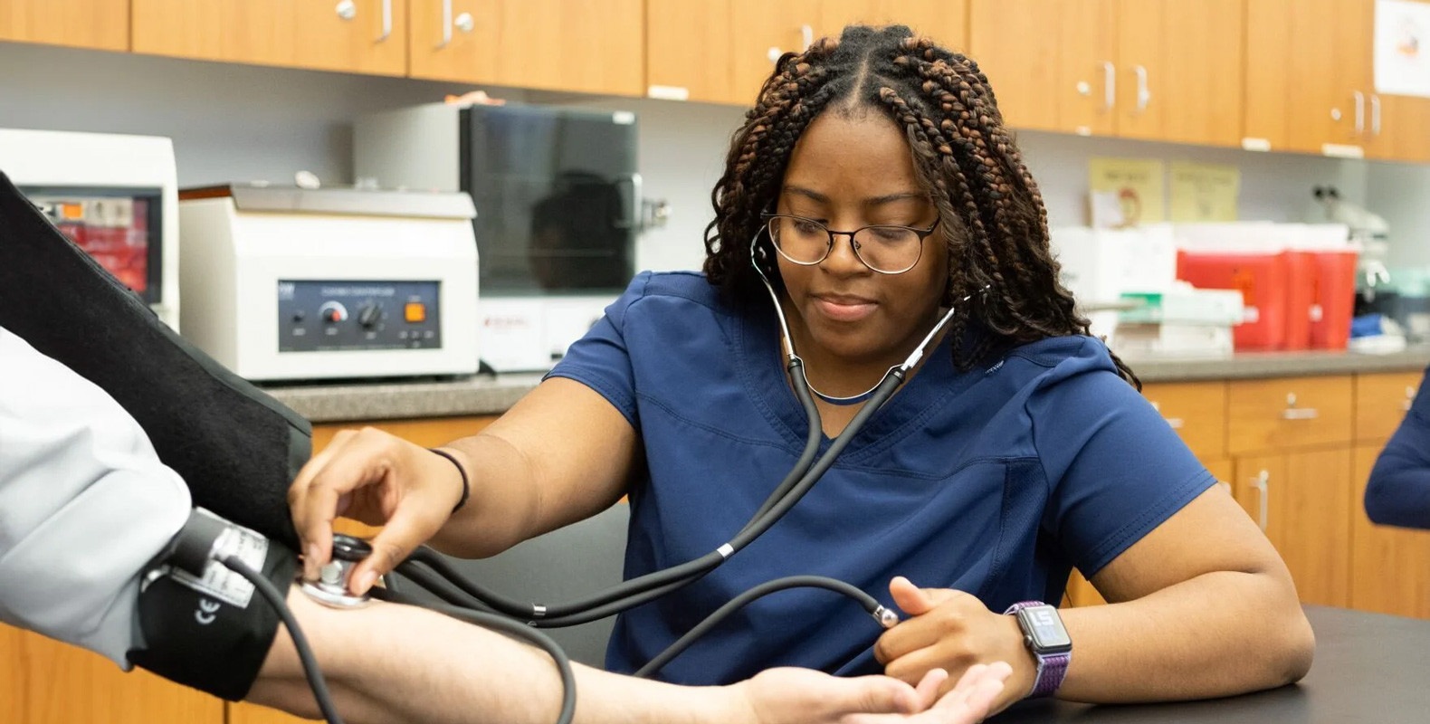 A healthcare worker in navy scrubs uses a stethoscope and blood pressure cuff to check a patient's blood pressure in a medical office, reflecting the hands-on skills taught through 21st century learning at Bryant & Stratton College.