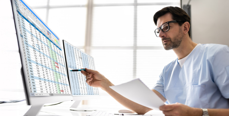 A man in medical scrubs and glasses sits at a desk, holding papers and pointing a pen at data displayed on two large computer monitors, which show spreadsheets with rows of numbers and colored cells.