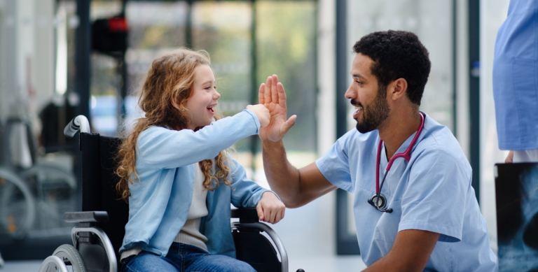 A smiling healthcare professional gives a high five to a happy young girl in a wheelchair inside a bright medical facility.