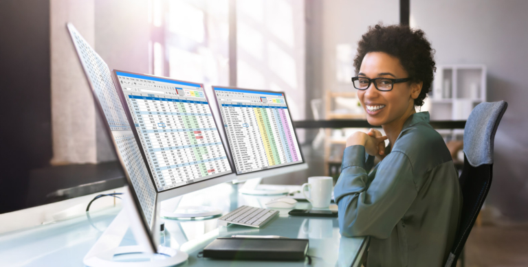 A woman wearing glasses sits at a desk, smiling at the camera, with two computer monitors displaying spreadsheets and data. Modern office setting with sunlight streaming through large windows.