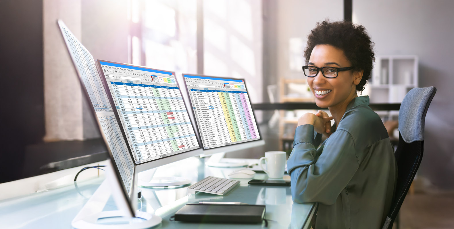 A woman wearing glasses sits at a desk, smiling at the camera, with two computer monitors displaying spreadsheets and data. Modern office setting with sunlight streaming through large windows.