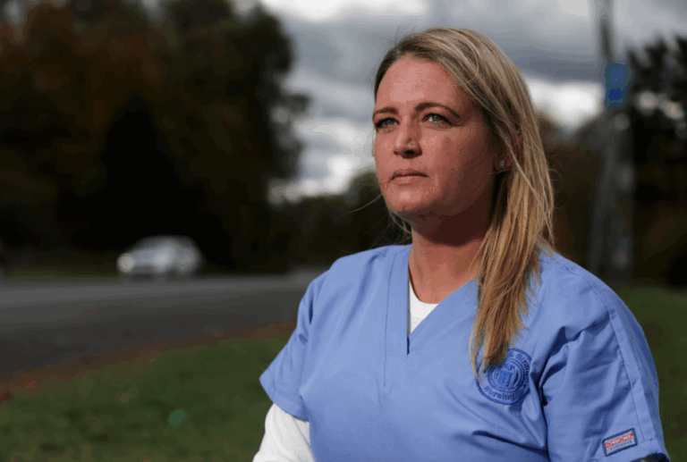 A woman in light blue medical scrubs stands outdoors near a road, looking thoughtfully into the distance. Trees and a cloudy sky are visible in the background, reflecting the spirit of Bryant & Stratton College’s commitment to online enrollment.