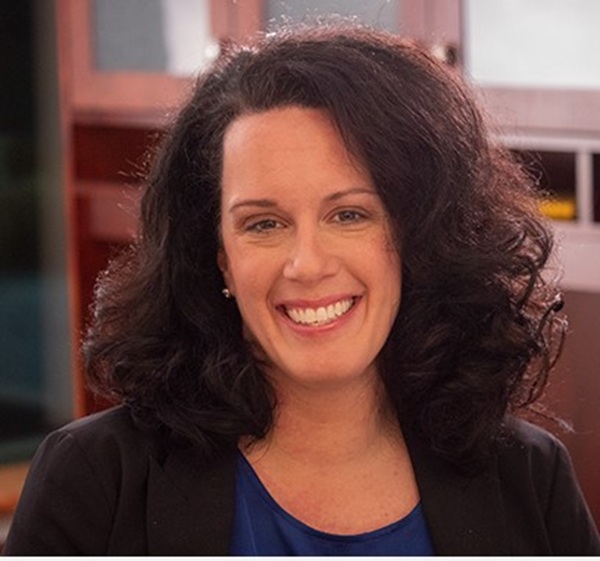 A woman with curly dark hair, wearing a black blazer and a blue top, is smiling at the camera in an indoor setting with blurred office furniture in the background, representing Bryant & Stratton College as a dedicated nursing student.