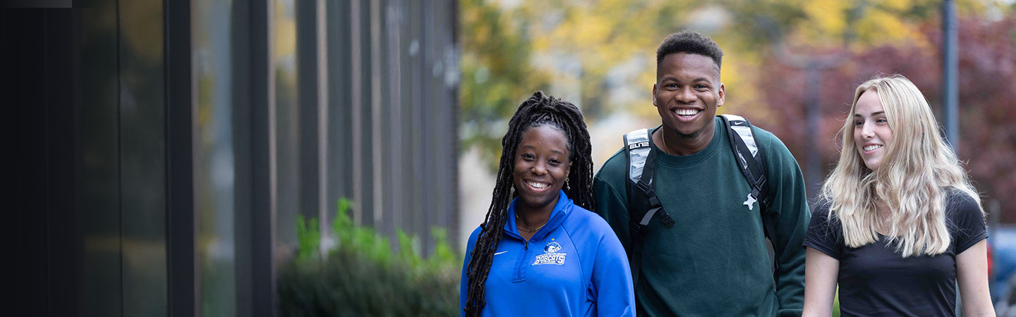 Three students, two women and one man, walk outdoors smiling on College Visit Day. They appear to be on a college campus, with trees and buildings in the background on a crisp fall day.