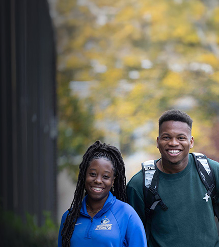 Two students smiling outdoors on College Visit Day, one in a blue shirt and the other in a dark green sweater with a backpack. Trees with yellow-green leaves are blurred in the background, creating a welcoming Information Hub atmosphere.