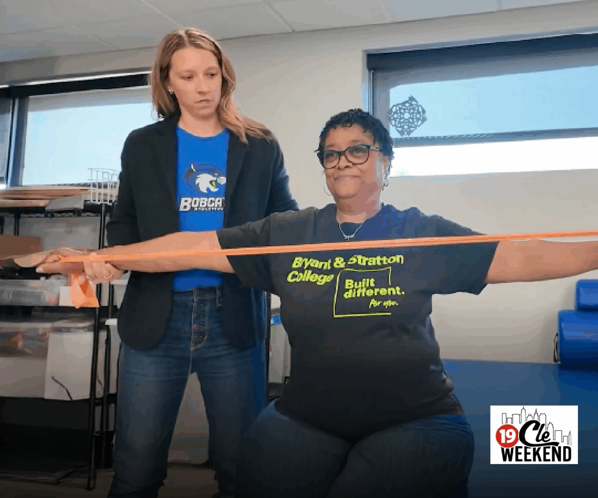 A woman in a Bryant & Stratton College shirt, representing student-athletes, does arm exercises with an orange resistance band, assisted by another woman in a blue Bobcats shirt. The CLE Weekend logo appears in the bottom-right corner.