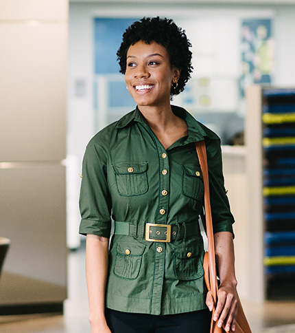 A woman with short curly hair, wearing a green belted button-up shirt and carrying a brown shoulder bag, smiles while standing indoors in a brightly lit, modern Racine office space during Registration Days.
