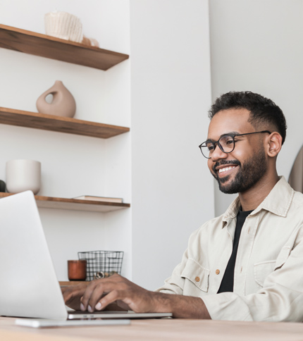 A man wearing glasses and a light-colored shirt is smiling while typing on a laptop at a desk, possibly drafting a Request Info email. Wooden shelves with decorative items are visible in the background.