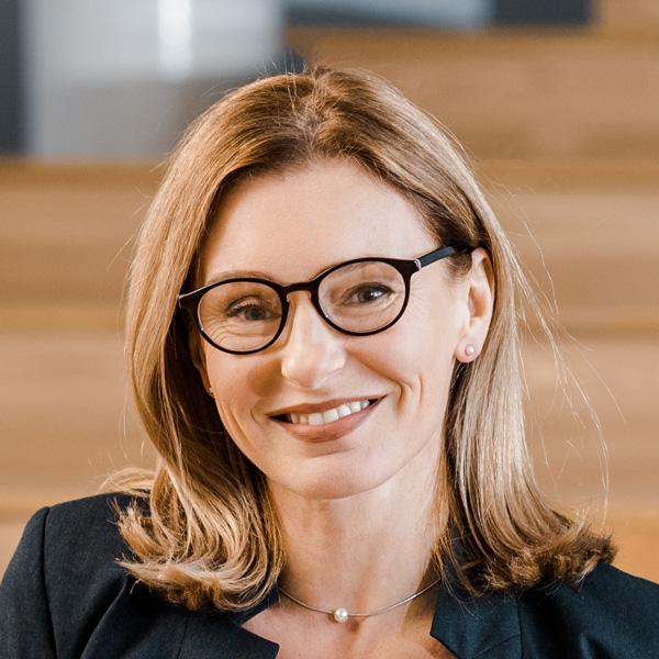 Sandra K. Whitmore, DNP, RN, with shoulder-length blonde hair, glasses, a dark blazer, and a necklace, smiles at the camera. She is sitting indoors with wooden steps in the background.
