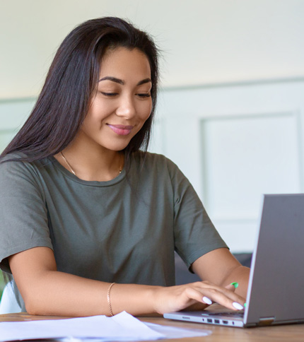 A young woman with long dark hair, wearing a gray t-shirt, smiles while typing on a laptop at a table with papers in front of her.
