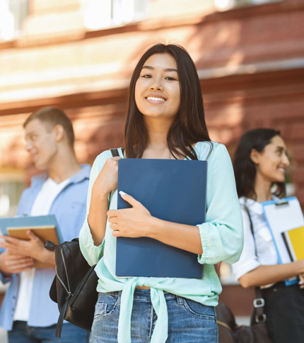 A young woman standing outdoors, smiling and holding a blue folder, with two other students in the background, in front of a brick building. All appear to be college students.