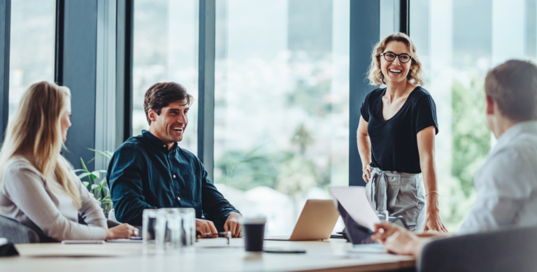 Four colleagues sit and stand around a conference table in a modern office, smiling and talking. Laptops, notebooks, and water glasses are on the table. Large windows show an out-of-focus cityscape in the background.