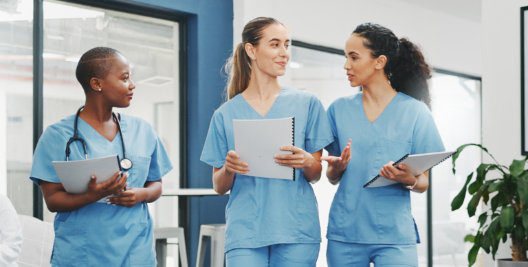 Three healthcare professionals in blue scrubs walk together in a hospital hallway, holding clipboards and notebooks, and discussing an upcoming CCNE accreditation review.