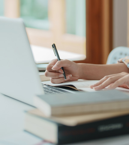 A person writes in a notebook with a pen while using a laptop, seated at a desk with books stacked nearby in a well-lit room, preparing notes about the SARA Student Complaint Process.
