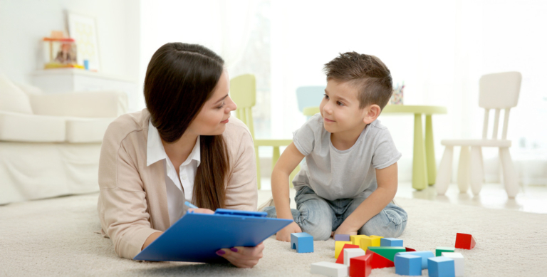A woman holding a blue clipboard lies on the floor facing a young boy who is sitting beside colorful toy blocks. They are indoors in a bright, child-friendly room, appearing to engage in conversation or an activity.